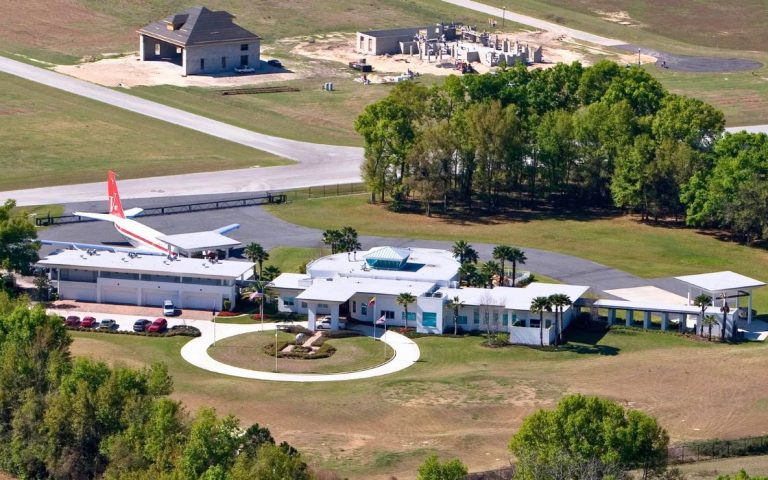 An aerial view of John Travolta's elegant home in Ocala, Florida, showcasing its architectural beauty amidst a serene landscape, complete with a private aircraft parked in front and a solitary tree standing tall in the field.